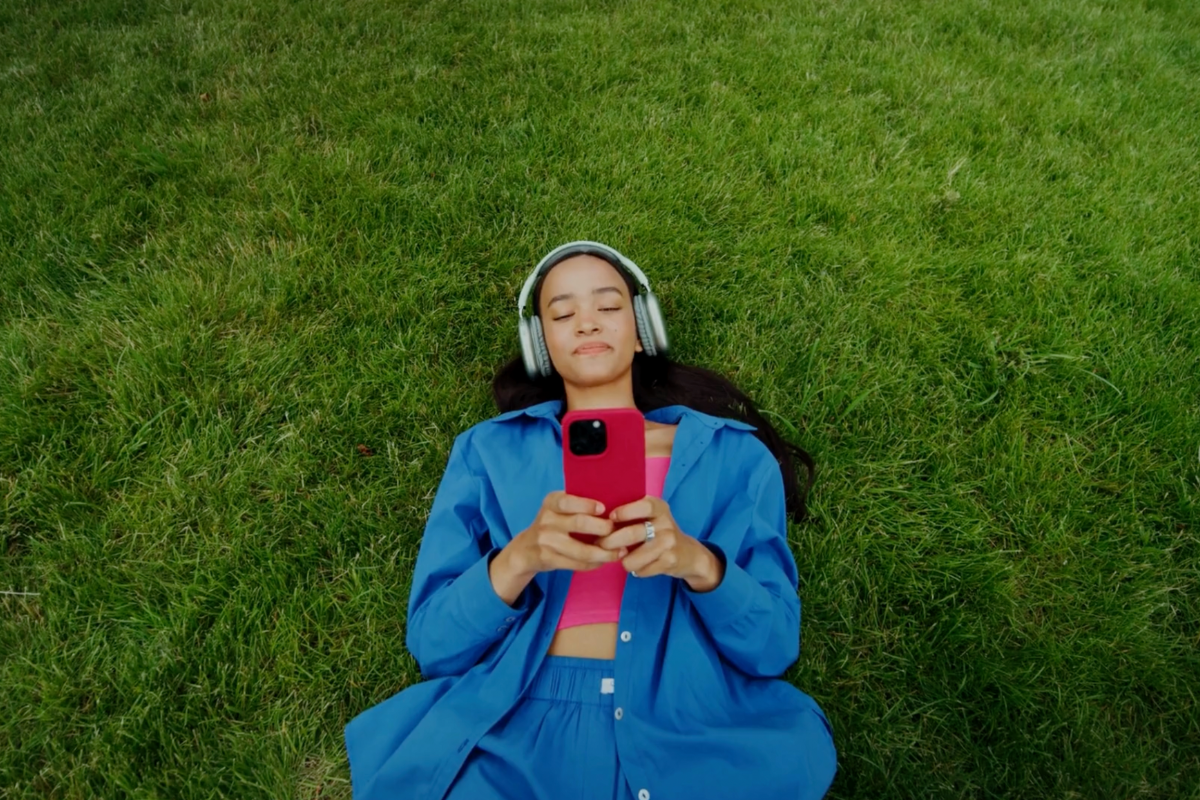 A young woman holding an iPhone with headphones relaxing on a green field.