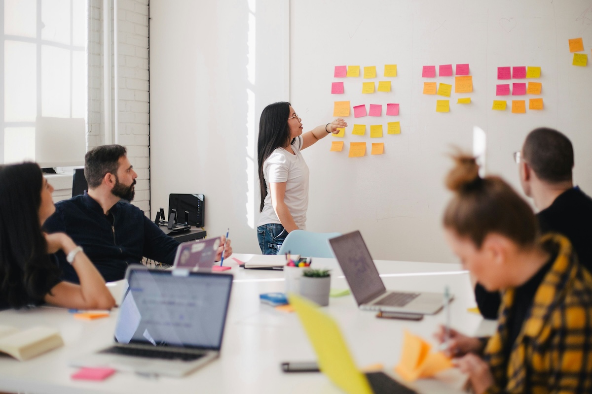 Woman presenting to colleagues in bright office, pointing at colorful sticky notes on wall. Team seated at table with laptops, engaged in brainstorming session