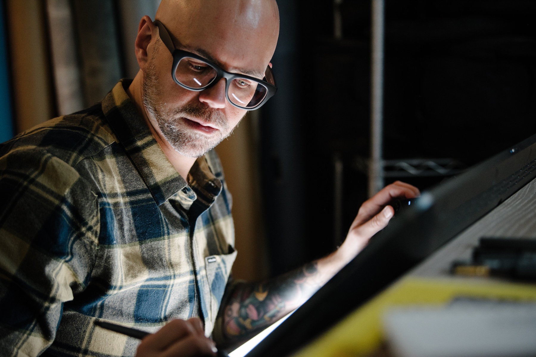 Person with glasses and plaid shirt looking at tablet or screen in dark setting.