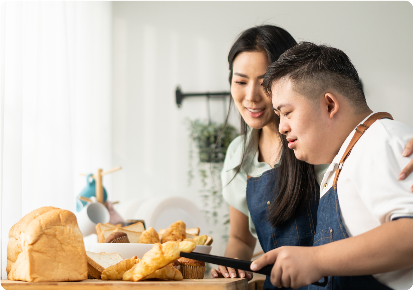 A woman and a man with Down syndrome wearing aprons stand together in a kitchen, smiling and looking at a tray with various bread items. The man holds a knife, ready to cut the bread.