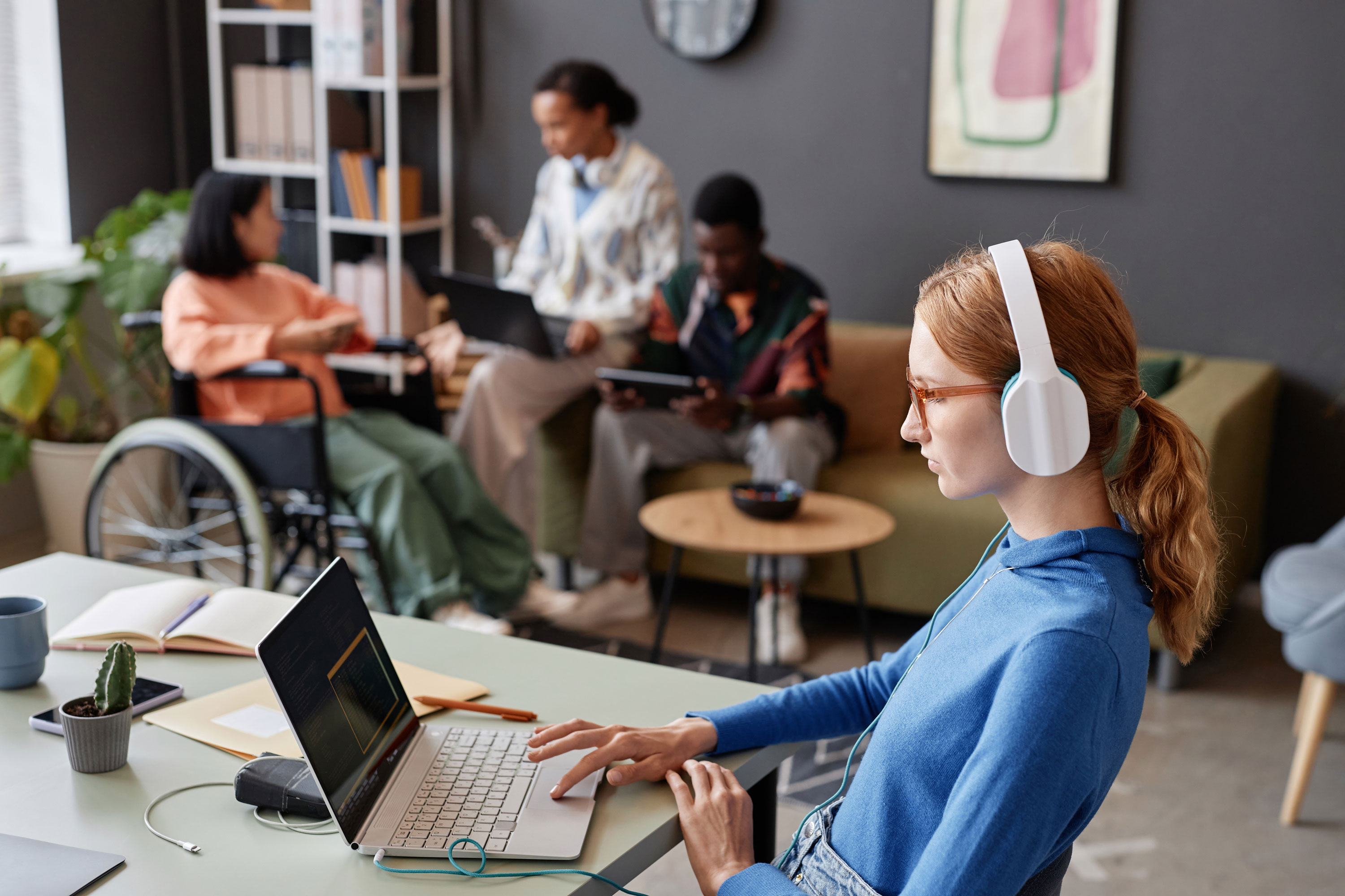 A woman wearing headphones sits at a desk with a laptop, while a person in a wheelchair chats with others in the background.