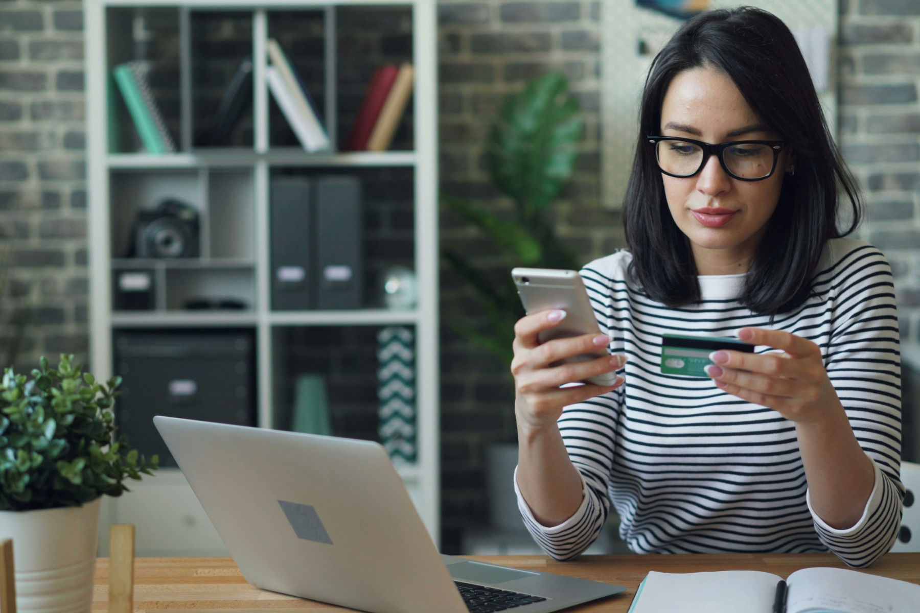 Woman using smartphone and credit card at desk with laptop.