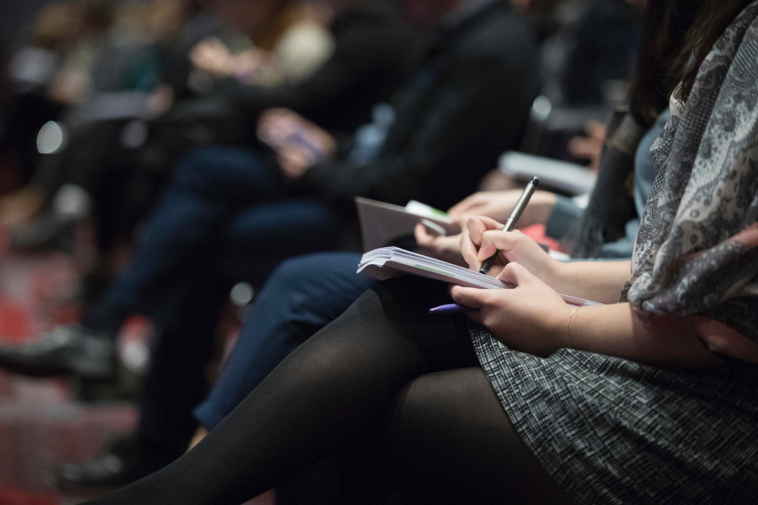 Person taking notes in auditorium or conference, focused on notebook and pen